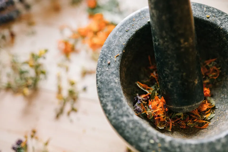 Nahaufnahme eines Steingefäßes mit Mörser und Stößel, der lebendige getrocknete Blumen auf einer Holzoberfläche mahlt.